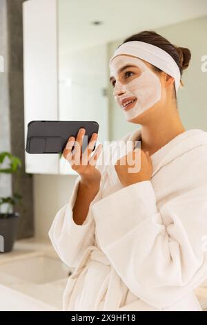 Femme caucasienne à la maison portant un masque facial, tenant un téléphone dans une salle de bain semblable à un spa. Elle porte un peignoir blanc, profitant d'un moment de détente et de détente Banque D'Images