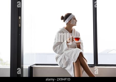 À la maison, une jeune femme caucasienne se détend avec un masque facial et du thé par une grande fenêtre, espace copie. Elle profite d'un moment de paix, prenant le temps de se désunw Banque D'Images
