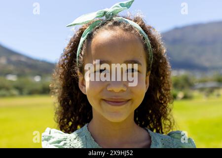 Fille biraciale avec des cheveux bouclés souriant à l'extérieur, bandeau vert assorti à sa robe Banque D'Images