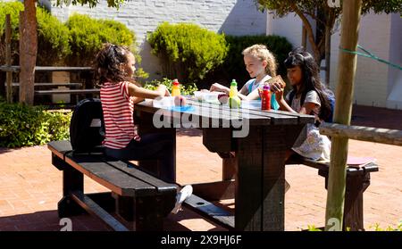 Trois enfants profitent d'un déjeuner ensoleillé en plein air à une table en bois à l'école Banque D'Images