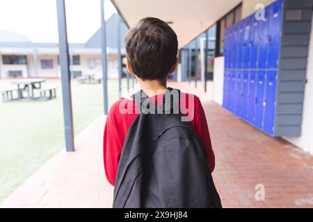 À l'école, jeune garçon biracial portant une chemise rouge et un sac à dos marchant à l'extérieur Banque D'Images