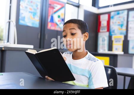 Biracial Boy se concentre sur la lecture dans une salle de classe ornée d'affiches éducatives. Banque D'Images