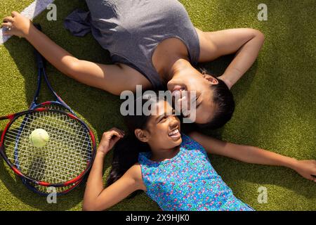 À l'extérieur, mère biraciale et fille allongées sur l'herbe avec des raquettes de tennis, souriantes Banque D'Images