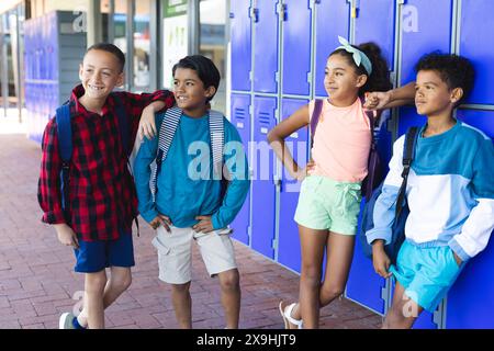 Trois garçons biraciaux et une fille biraciale se tiennent près de casiers à l'école, souriant Banque D'Images