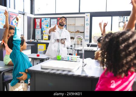 Jeune enseignant asiatique enseignant une classe de sciences à l'école, divers enfants levant les mains avec impatience. Filles biraciales et garçon engagés dans l'apprentissage, la classe Banque D'Images