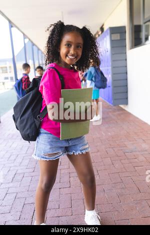 Fille biraciale aux cheveux bouclés souriant, tenant un dossier vert et portant une chemise rose à l'école Banque D'Images