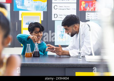 À l'école, enseignant asiatique guidant un étudiant biracial dans une expérience scientifique. Les deux examinent des liquides colorés dans des flacons, entourés b Banque D'Images