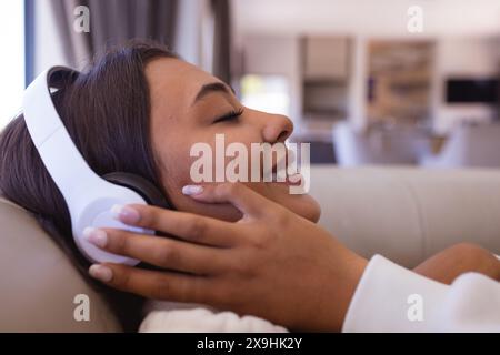 Une jeune femme biraciale portant des écouteurs blancs, se relaxant à la maison Banque D'Images