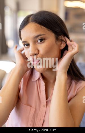 Une jeune fille biraciale ajustant sa boucle d'oreille à la maison, portant une chemise rose Banque D'Images