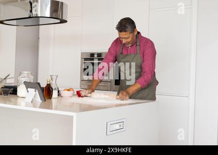 À la maison, homme senior biracial avec les cheveux gris et la barbe en chemise rouge et tablier gris. Rouler la pâte et regarder le comprimé sur le comptoir pendant la cuisson, unalt Banque D'Images