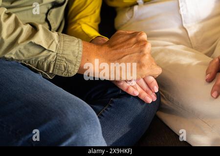 À la maison, un couple de personnes âgées diversifié est assis étroitement sur un canapé, se tenant les mains doucement. L'un portant une chemise verte et un Jean, l'autre dans une chemise jaune et light-c Banque D'Images