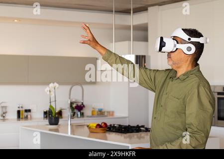 À la maison, homme senior biracial dans la cuisine moderne portant un casque de réalité virtuelle, espace de copie. Tendre la main, interagir avec envi virtuel Banque D'Images