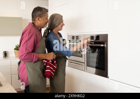 À la maison, couple senior diversifié utilisant le four, à la fois portant des tabliers et souriant. Mari biracial d'âge moyen avec les cheveux gris et femme caucasienne tenant le kit Banque D'Images