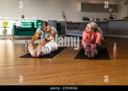 À la maison, couple senior diversifié s'étirant sur des tapis de yoga dans un salon lumineux. Homme biracial avec les cheveux bruns courts et femme caucasienne avec long hai gris Banque D'Images