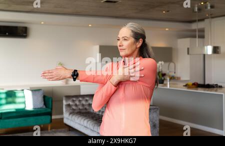 À la maison, une femme âgée caucasienne aux longs cheveux gris s'étire dans un salon moderne. Portant un haut rose à manches longues et une smartwatch noire, look Focu Banque D'Images