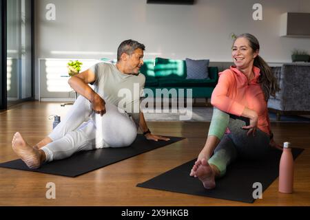 À la maison, couple senior diversifié pratiquant le yoga ensemble sur des tapis dans le salon. Homme biracial avec les cheveux gris courts et femme caucasienne avec long h gris Banque D'Images