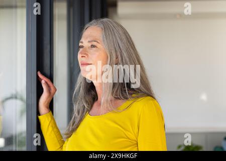 Femme âgée caucasienne avec de longs cheveux gris et des yeux bleus debout près de la fenêtre à la maison. Portant une chemise jaune, regardant dehors réfléchi avec sa main Banque D'Images