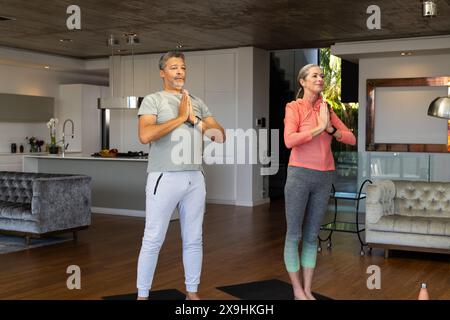 À la maison, couple senior diversifié pratiquant le yoga dans un salon moderne. Femme caucasienne mature avec les cheveux gris en queue de cheval et homme biracial senior avec s. Banque D'Images