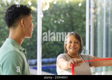 À la maison, un jeune physiothérapeute masculin biracial aide une patiente biraciale senior. Ils sont dans une chambre lumineuse et spacieuse avec de grandes fenêtres donnant sur Banque D'Images