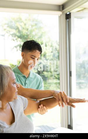 Le jeune thérapeute masculin biracial aide les patientes biraciales âgées à faire de l'exercice à la maison. Ils sont dans une chambre lumineuse et ensoleillée avec de grandes fenêtres donnant sur un Banque D'Images