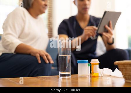 À la maison, un jeune infirmier biracial montre un comprimé et un médicament à une patiente âgée. Ils sont dans un cabinet médical avec des bouteilles de médicaments et Banque D'Images