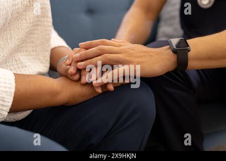 Jeune infirmière masculine biraciale aide la patiente senior à la maison se tenant la main. Ils sont assis sur le canapé bleu dans un environnement confortable et favorable, un Banque D'Images