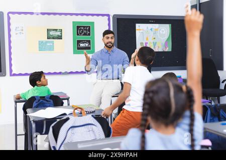 Jeune enseignant asiatique enseignant à un groupe diversifié d'enfants dans une salle de classe. Il s'engage avec les étudiants, qui lèvent la main pour répondre Banque D'Images