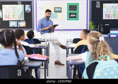 Jeune enseignant s'engage avec un groupe diversifié d'élèves dans une salle de classe d'école colorée. Il est assis sur un bureau avec une tablette à la main, favorisant un interacti Banque D'Images