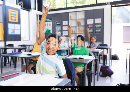 Garçon biracial lève la main avec impatience dans une salle de classe. D'autres enfants, y compris une fille biraciale, sont également désireux de participer à la leçon. Banque D'Images