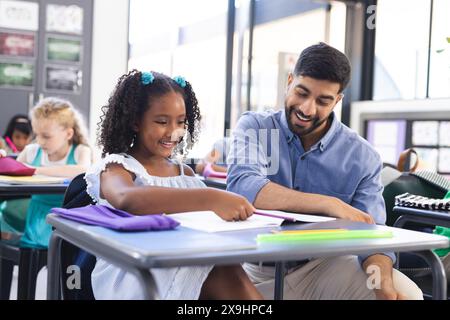 À l'école, enseignant asiatique aidant de jeunes étudiantes biraciales en classe. Les deux sont assis à un bureau avec des fournitures scolaires, souriant comme ils foc Banque D'Images