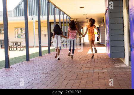 Trois filles biraciales se tiennent la main et courent dans un couloir scolaire avec un espace de copie Banque D'Images