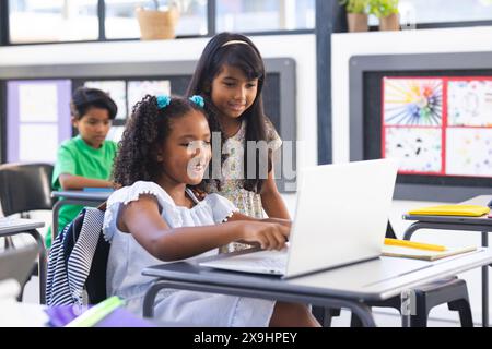 Deux filles biraciales, une en robe blanche, utilisent un ordinateur portable dans une salle de classe Banque D'Images