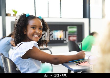 Fille biraciale avec les cheveux bouclés sourit tout en tenant un crayon dans une classe d'école Banque D'Images