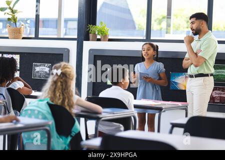 À l'école, un jeune enseignant asiatique écoute une fille biraciale présenter Banque D'Images