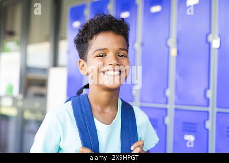 Garçon biracial aux cheveux bouclés sourit devant des casiers bleus à l'école, portant un sac à dos Banque D'Images