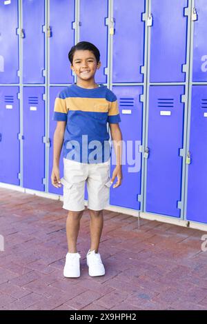 Garçon biracial se tient souriant devant des casiers bleus à l'école, habillé de façon décontractée Banque D'Images