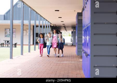 Quatre jeunes filles biraciales marchent ensemble dehors à l'école dans des vêtements décontractés colorés Banque D'Images