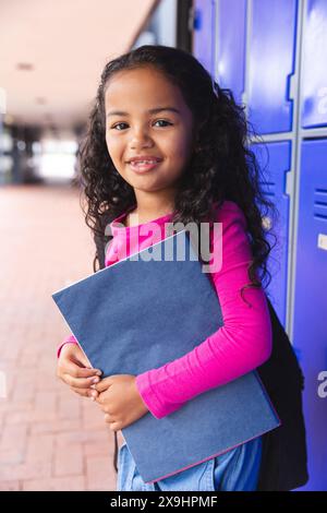 À l'école, à l'extérieur, une jeune fille biraciale tenant un dossier bleu se tient près de casiers Banque D'Images