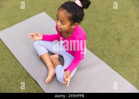 Une jeune fille biraciale portant du rose est assise sur un tapis de yoga à l'extérieur Banque D'Images