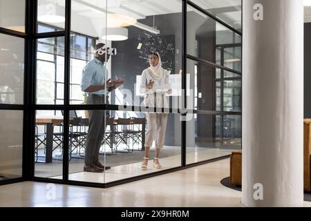 Dans le bureau d'affaires moderne, équipe diversifiée discutant des dessins d'architecte sur le mur de verre. Jeune homme afro-américain en chemise bleue et lunettes, et jeune Banque D'Images