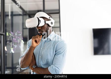 Jeune homme afro-américain portant un casque de réalité virtuelle dans un bureau d'affaires moderne. Il est attentionné, avec des cheveux noirs courts, barbe taillée, et l'usure Banque D'Images