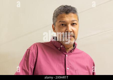 Homme senior biracial avec les cheveux gris courts et la barbe taillée dans une chemise rose à la maison, regardant la caméra. Il a une expression réfléchie, debout à nouveau Banque D'Images