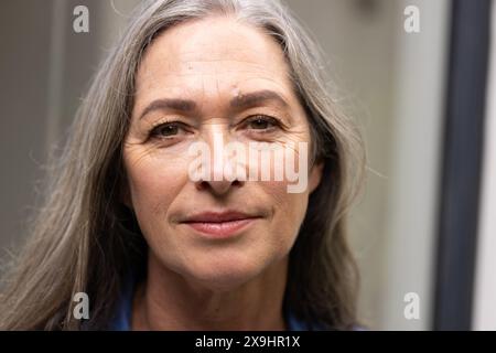 À la maison, femme caucasienne senior avec de longs cheveux gris et des rides sur son visage. Peau claire, yeux bruns, portant une chemise bleue, regardant directement la caméra Banque D'Images