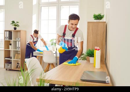 Joyeux hommes jeunes nettoyeurs professionnels essuyant le bureau dans le bureau avec des fournitures de détergent ménager. Banque D'Images