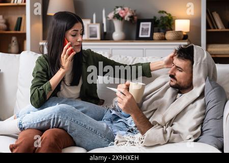 Jeune femme aidant et traite son homme malade à la maison qui est enveloppé dans une couverture avec une température corporelle élevée. Homme malade du rhume et de la grippe mesure TEM Banque D'Images