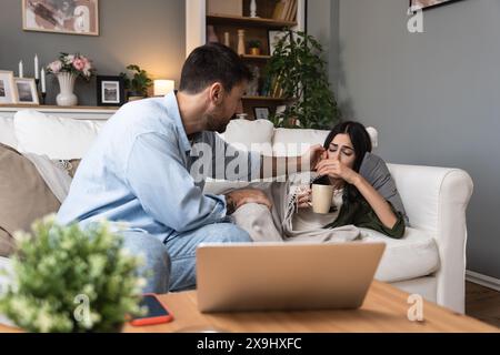 Jeune homme aidant et traite sa femme malade à la maison qui est enveloppée dans une couverture avec une température corporelle élevée. Femelle malade du rhume et de la grippe mesurant t Banque D'Images