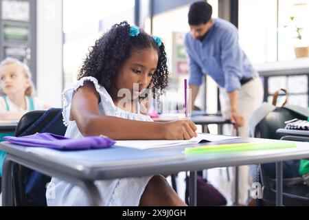 À l'école, jeune fille biraciale se concentrant sur l'écriture dans la salle de classe, enseignant asiatique aidant. Elle a les cheveux bouclés avec des noeuds bleus, il porte un blu Banque D'Images