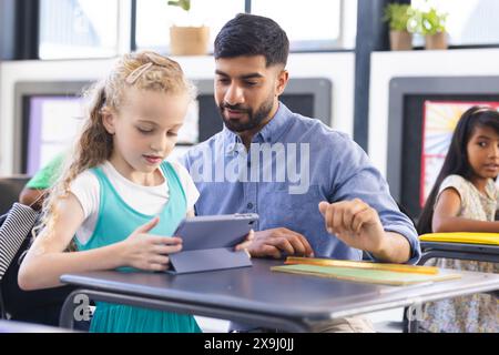 À l'école, enseignant asiatique montrant tablette à une jeune fille caucasienne en classe. Fille biraciale regardant, jeune garçon biracial lisant à proximité, inaltérée Banque D'Images