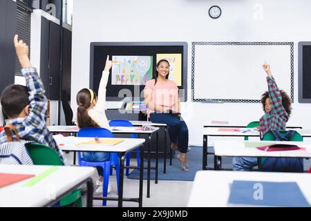 À l'école, une jeune enseignante biraciale sourit aux élèves levant la main en classe Banque D'Images