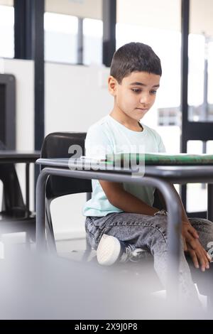 À l'école, jeune étudiant biracial assis à un bureau dans une salle de classe, regardant une tablette Banque D'Images